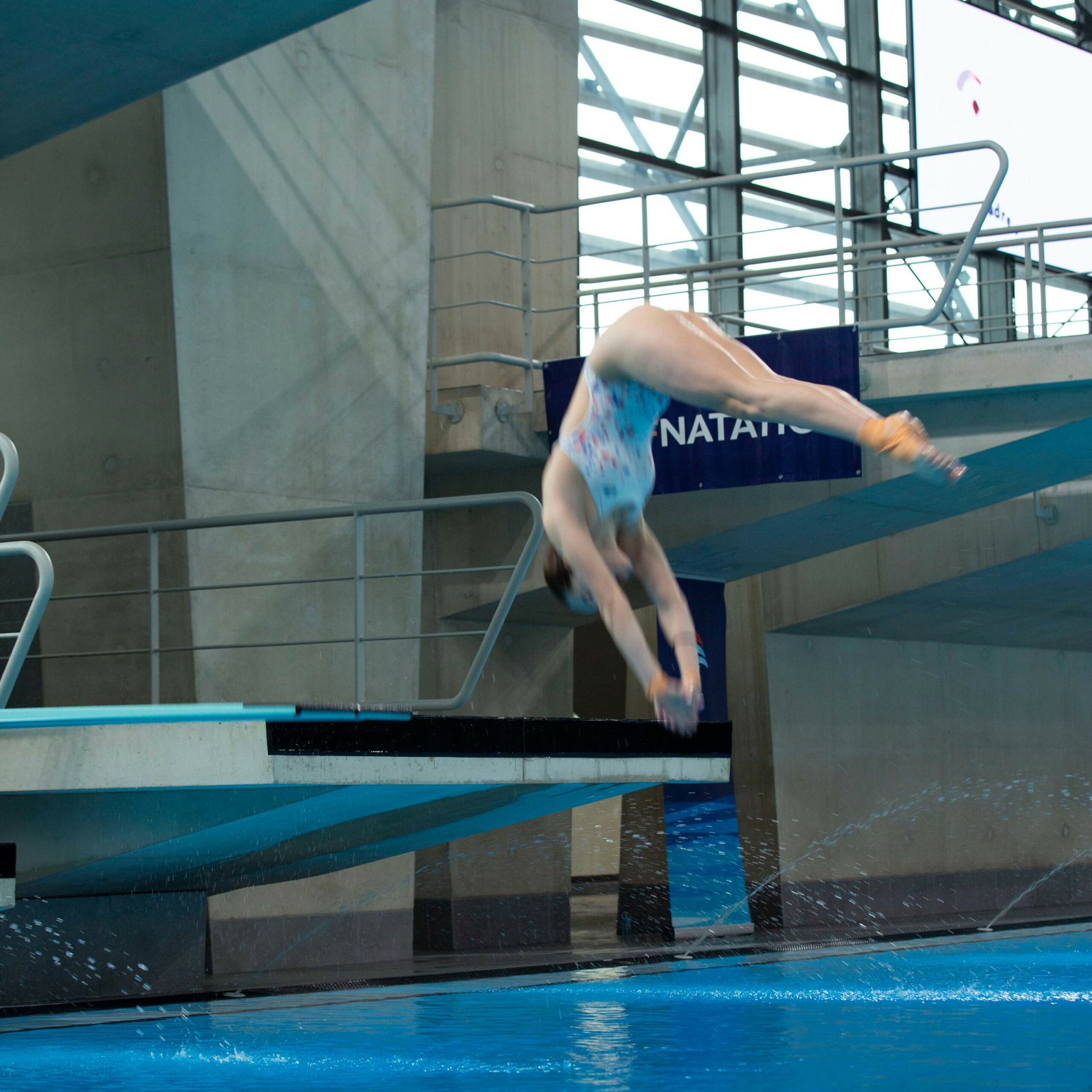 Plongeuse effectuant un plongeon retourné au tremplin de 1 m au Centre Aquatique Olympique Métropole du Grand Paris